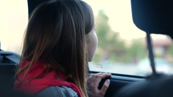 Close up of Caucasian pretty girl looking through car window on street with amazed face pointing at something talking to mom. Little teen kid sitting in automobile looks out the window. Trip. Travel