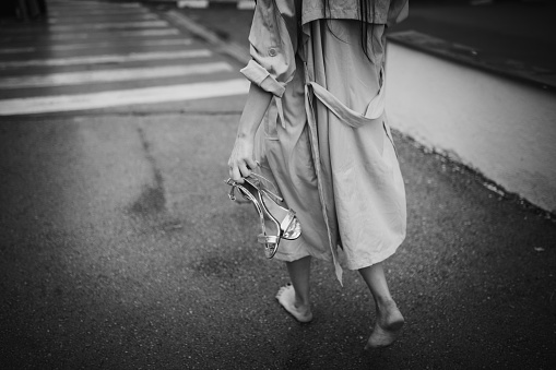 A young beautiful woman is walking down the street after a night out. She is holding heels in her hands and the street is wet. Unrecognizable person.