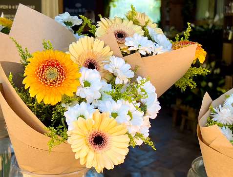 Horizontal closeup photo of white chrysanthemums and yellow gerbera wrapped simply in brown paper, outside a Florist shop. Byron Bay, NSW.