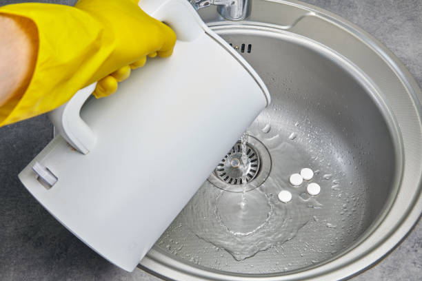 a person in yellow rubber gloves pours hot water with steam from an electric kettle into a metal sink - sink drain plumber domestic kitchen imagens e fotografias de stock