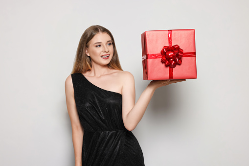 Portrait of beautiful young woman in elegant black dress with red gift box on white background