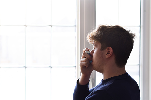 Closeup side view young man standing in front of a window using puffer inhaler to control his asthma