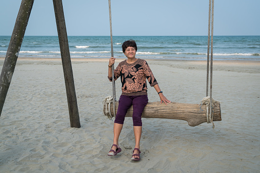 Senior Asian woman relaxing on a wood plank swing on the beach. Retirement or travel vacation concept.