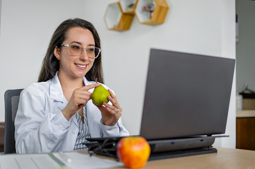 A Latina nutritionist sitting in her office in an online video call with a patient who she recommends eating fresh fruit.