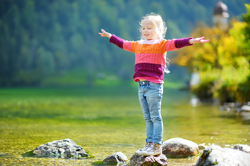 Adorable girl playing by Konigssee lake in Germany on warm summer day. Cute child having fun feeding ducks and throwing stones into the lake. Summer activities for kids.