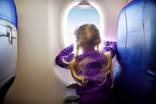 Adorable little girl traveling by an airplane. Child sitting by aircraft window and looking outside. Traveling with kids abroad.
