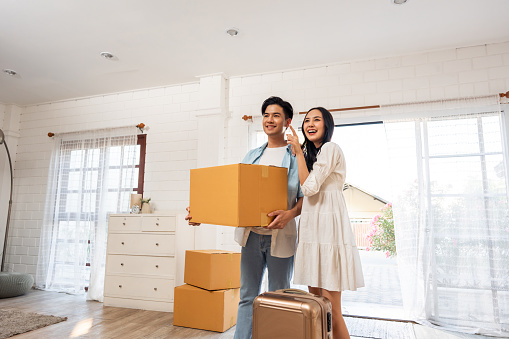 Asian young happy new marriage couple moving to new house together. Attractive romantic man and woman holding box parcel and suitcase with happiness and love. Family-Moving house relocation concept.