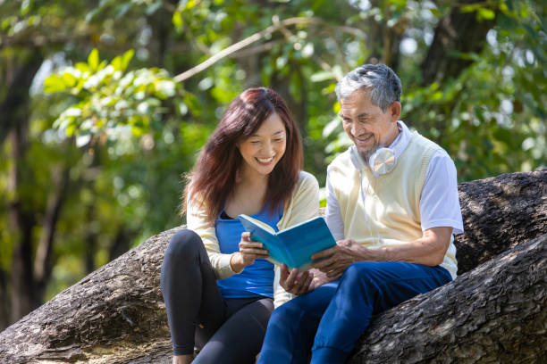アジアの年配の父と娘は、夏の間、成熟した木の枝に座って一緒に本を読んで、心身の健康と長寿の使用を目的としています。 - grandparent senior adult child reading ストックフォトと画像