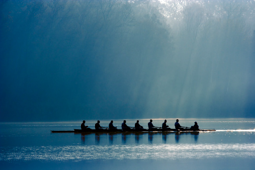 A canoe team rowing with power in order to win the competition. On background morning light through the forest. 