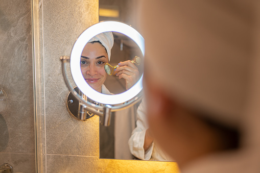 Woman in Bathrobe Using Jade Roller in Front of Mirror in Bathroom