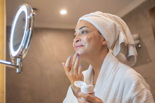 Woman in Bathrobe Applying Cream to Her Face in the Bathroom