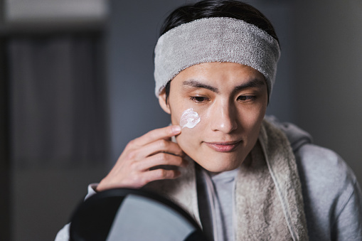 A Japanese man in his thirties (men's beauty) who paints skin care cream on his face while looking in the mirror at home