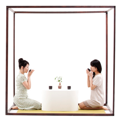 Two Japanese woman ,kneeling within a wood framed cube, drinking tea. 