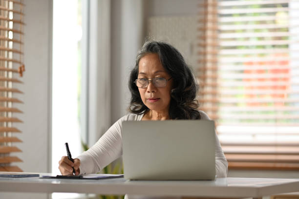 istock Smiling senior woman in glasses using laptop and making notes on paper. People, technology and remote work concept. 1485028808