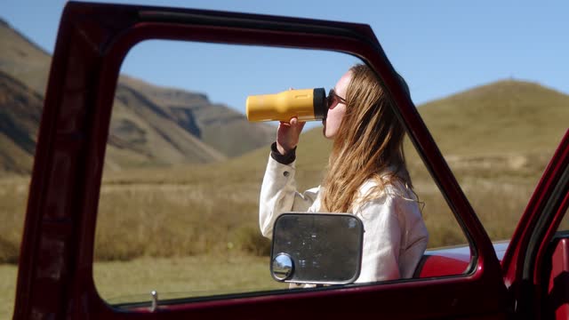 Attractive long-haired girl drinking a drink from a thermos of thermo mugs while standing near a vintage car against the backdrop of a mountain landscape.