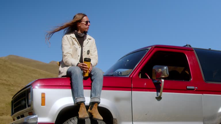 Attractive long-haired girl drinking a drink from a thermos of thermo mugs while standing near a vintage car against the backdrop of a mountain landscape.