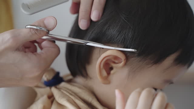 Young Asian mother cutting hair to his baby boy by herself at home. Smiling little cute Asian baby boy getting his first haircut by his mom. Asian family urban lifestyle at home, parent child relation concept.