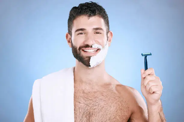 Portrait of happy man with foam on beard, razor and towel in skin product placement in studio mock up. Shaving cream on face, smile and hair or skincare for male model, isolated on blue background.