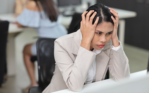Adult asian businesswoman sitting and touching her head and looking at computer in office after bad news business failure or get fired and feeling discouraged, distraught and hopeless in modern office