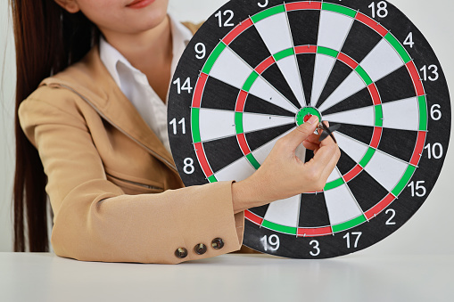Businesswoman hands in classic suite sitting and holding black dart put to centre of target board on white table meaning of investment and achievement goal. Planning and strategy concept.