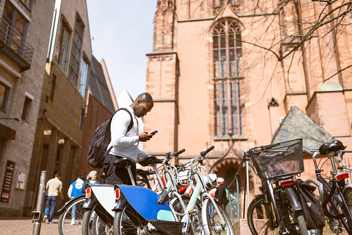 black tourist in Frankfurt renting a city bike through a mobile application. He is  is casually dressed and enjoying a bike ride while exploring the beautiful streets and landmarks of the city. This image represents a blend of adventure, diversity, and exploration, highlighting the joy of discovering a new destination.