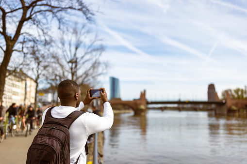 Black tourist in Frankfurt, casually strolling along the banks of the Main River. He is dressed in casual attire and carries a backpack. Engrossed in the beauty of his surroundings, with a bridge visible in the background, he captures beautiful Main River with his camera.