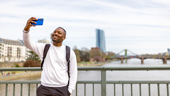 Black tourist in Frankfurt, casually strolling along the banks of the Main River. He is dressed in casual attire and carries a backpack. Engrossed in the beauty of his surroundings, with a bridge visible in the background, he taking selfie.