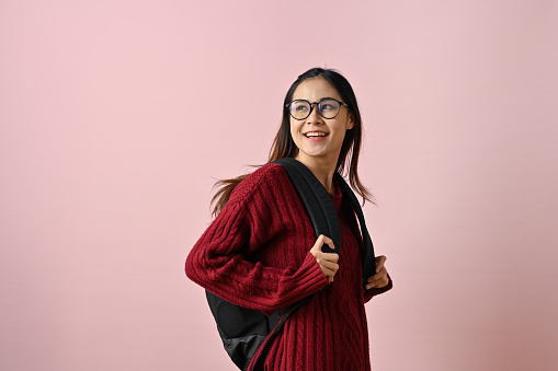 Pretty and wise young Asian female college student in a red knitted sweater and glasses stands against an isolated pink background with her backpack.