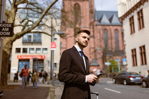 A poised young businessman, dressed in a sharp suit, is captured on a business trip in Frankfurt. With confidence, he holds a small carry-on suitcase, ready for travel. Engrossed in a phone conversation, he efficiently handles important matters while on the move. The vibrant cityscape sets the backdrop, reflecting the dynamic and modern business environment.
