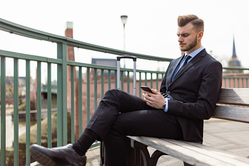 Young business professional sitting on a bench in Frankfurt, Germany, while using his mobile phone. He is elegantly dressed and carries a briefcase, embodying a professional demeanor.