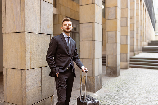 Young businessman on a business trip in Frankfurt, Germany. He is exuding elegance with his well-groomed appearance and professional attire. The young man is seen holding a sleek, black briefcase, symbolizing his preparedness and dedication to his work.