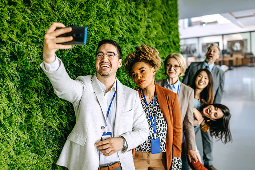 Colleagues taking selfie at the office, standing near green wall with plants
