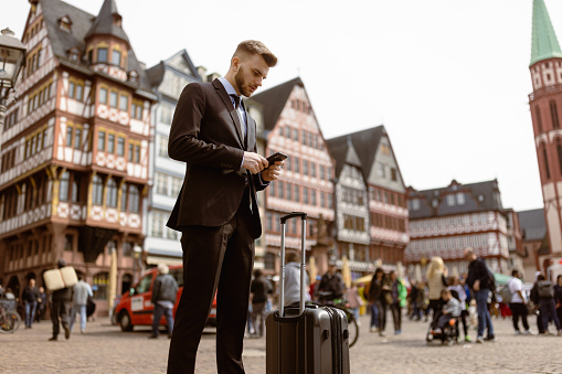 Successful young man in a business suit, who is currently on a business trip in Frankfurt. He stands at Romer Square, a historic and bustling location in the city. With a confident demeanor, he holds a small carry-on suitcase, indicating his readiness for travel. Engrossed in a phone conversation, it's evident that he is a busy professional, handling important matters even while on the move.