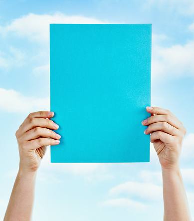 A woman's hands hold up an A4-sized sheet of blank, blue paper - ready for your message - against a lightly cloudy sky.