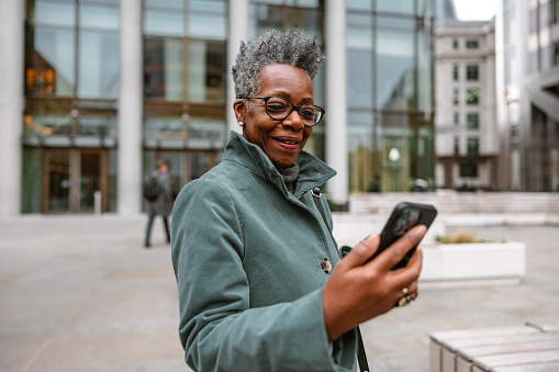 A happy senior adult black woman using a smartphone outdoors. She is standing outside on a city street while looking at the phone screen. The woman is wearing an elegant coat. The weather is gloomy and cold. She is smiling  and looks relaxed.