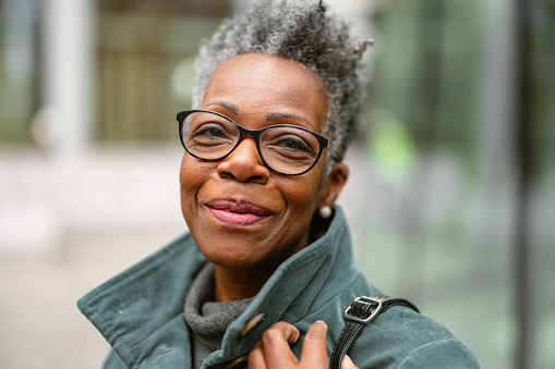A portrait of a senior adult black female outdoors looking at the camera. She is smiling and looks happy. The female has short grey hair and is wearing glasses. The weather is gloomy and cold.