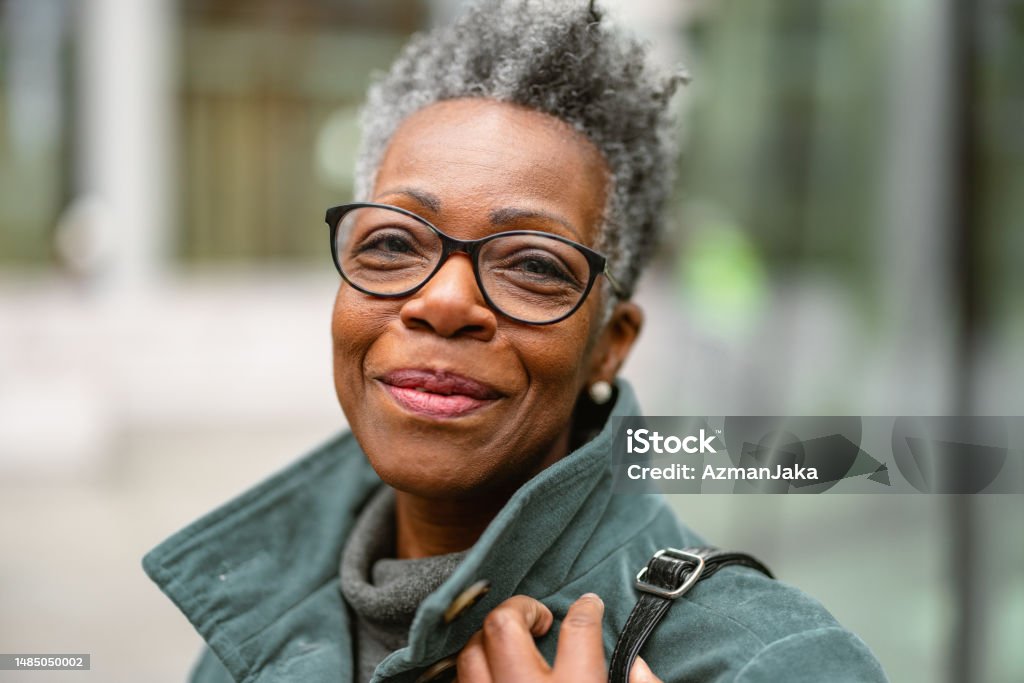 Portrait Of A Senior Adult Black Female On A City Street Looking AT The Camera A portrait of a senior adult black female outdoors looking at the camera. She is smiling and looks happy. The female has short grey hair and is wearing glasses. The weather is gloomy and cold. Portrait Stock Photo
