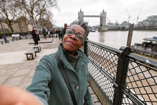 A senior adult black female tourist taking a selfie by the Tower Bridge to send to her children. She is visiting London alone during wintertime. The woman is standing by the fence on the riverbank of the river Thames. She looks happy to be exploring the city.