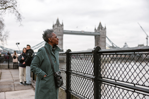 A senior adult black female pensioner admiring the river Thames during her daily stroll around London. The weather is cold and gloomy, so the woman is wearing a warm winter coat. In the background, there is a view of the famous Tower Bridge.