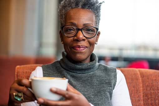 A senior adult black female smiling at the camera while holding a cup of delicious coffee in her hands. She is located in a cozy cafe in London on a gloomy winter day. The woman looks relaxed and content. She is dressed in cosy winter clothes and is wearing glasses.
