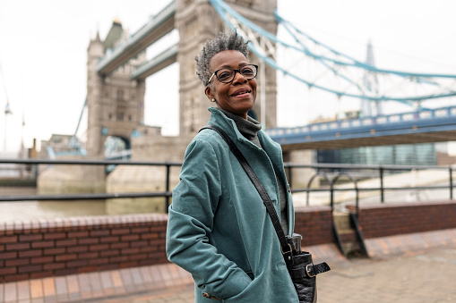 A senior adult black female tourist smiling at the camera while standing by the Tower Bridge in London on a gloomy winter day. She looks happy to be visiting London and exploring the city alone. The woman is wearing warm winter clothes.