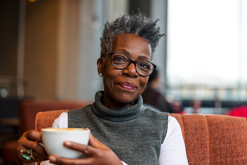 A senior adult black female looking at the camera while enjoying her relaxing morning at a cafe in London. She is holding a cup of coffee and smiling. The woman looks relaxed and calm. The weather outside is gloomy and cold.