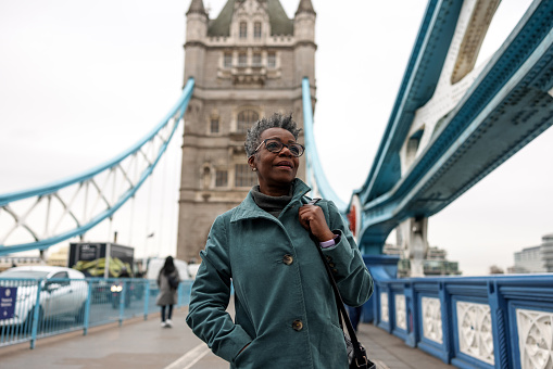 A senior adult black female pensioner strolling around London on a gloomy winter morning. She is looking around and observing her surroundings. The sky is grey and cloudy and the weather is cold. She looks calm and relaxed and is enjoying her morning walk.