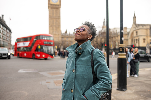 A senior adult black female tourist admiring the architecture in London. She is traveling alone and walking around the City of Westminster on a gloomy winter day. The woman is standing on the sidewalk and behind her, there is the famous Big Ben. She is looking up.