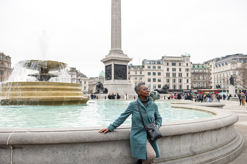 A senior adult black female tourist leaning on a fountain in London. She is located on the big Trafalgar square and looking around admiring the city. She is taking a break after a long day of visiting landmarks in London. The weather is cold and gloomy.