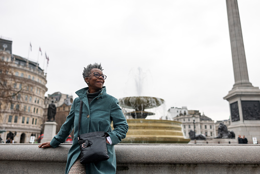 A senior adult black female tourist looking around while taking a break from sightseeing in London. She is located by the fountains at Trafalgar square. She is smiling and is happy to have the chance to explore London. The weather is cloudy and cold.