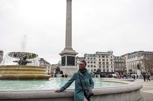 A senior adult black female tourist leaning on the fountain at the Trafalgar Square in London. She is taking a break to rest after a long stroll around the city. She is calm and relaxed. The woman is looking around and admiring the beautiful city.