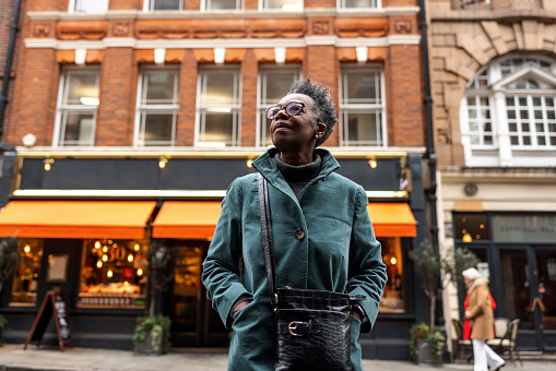 A senior adult black female tourist stopping on a street in London To admire the beautiful architecture in the city. She is located on a street with beautiful British buildings. The weather is cold and cloudy. The woman looks fascinated.