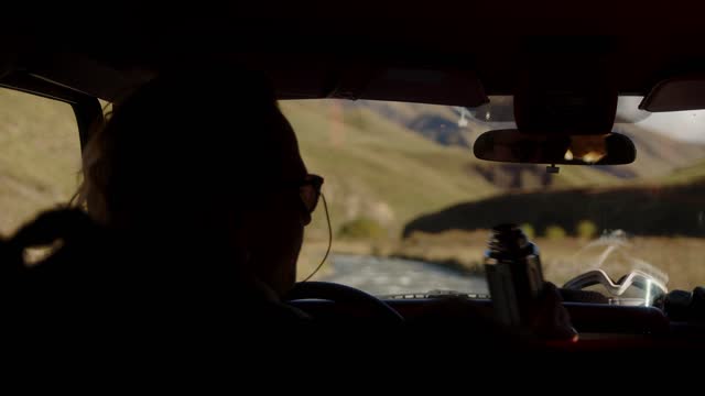 A man sits behind the wheel of an SUV and drinks hot coffee from a thermos. Traveling by off-road vehicle in the mountains. View from the cab of an SUV to the mountains and a mountain river.