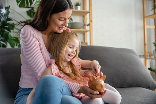 Cute little girl with her mother at home putting a coin into the piggy bank. Child savings plan concept